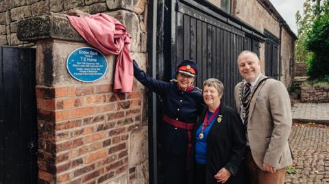 Deputy Lieutenant Judy Scott Moncrieff with Staffordshire Moorlands District Council chairman Adam Parkes and Moorlands Partnership Board chair Christina Jebb. The three are stood next a blue plaque on a brick wall, with Mrs Scott Moncrieff lifting a red curtain to reveal it.