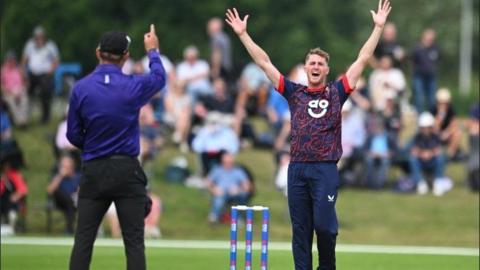 A Lancashire player in short-sleeved blue top with red facings and blue trousers appeals fgor a wciket while the umpire in bright blue shirt and black trousers wit h a black cap raises his right index finger to signify a dismissal. The stumps are blue with sponsor logo. A blurred shot of spectators sitting on a grass verge can be seen in the back ground.