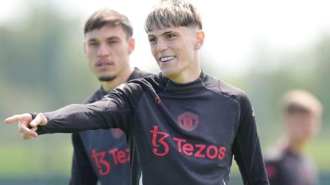 Garnacho has short dyed blonde hair and is wearing a black training top with red sponsor logo and Manchester United badge - it is a shot from training in 2025.