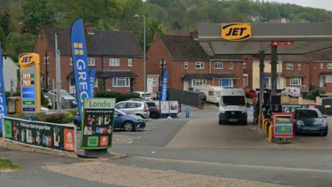 A Google Maps street view image of the entrance to a Jet petrol station. There are cars parked in bays to the left of the image and cars parked next to the petrol pumps. Three Greggs banners can also be seen to the left of the image.