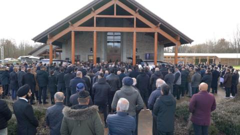 Crowds gathered for the funeral of Rajinder Verma. We see the backs of many people, mainly men, outside a building with two hearses just in front of the building. More than a hundred people can be seen. Many wear coats and some have head coverings. The building is made of stone with a roof which extends over the front, supported by wooden beams. One of the hearses has the boot open. 