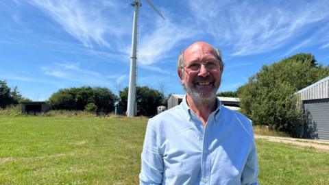 Pete West in a blue shirt in a field with a wind turbine in the background.