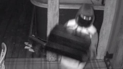 A black and white CCTV still of a person with their hood up while wearing a mask and carrying a black till. They are inside the pub, which has a wooden floor and wooden beams.