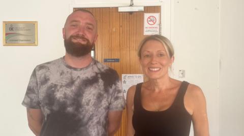 A man and woman stand indoors beside a wooden door covered with signs, including a No Smoking warning. The man on the left of the image is wearing a tie-dye t-shirt. The woman on the right is wearing a black tank top and light shorts. They are both smiling. A grey sofa and white walls frame the background.