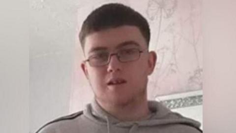 Daniel Ambler, a 23-year-old man with short dark hair, wearing spectacles and a grey hoodie. He is standing in what looks like a living room. There is pink and grey wallpaper on the wall behind him.