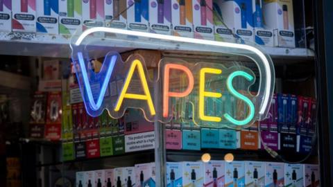 A brightly-lit sign reading "vapes" in a shop window 