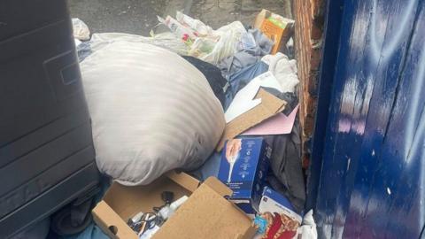 A pile of rubbish behind a large black commercial bin and in front of a blue, wooden back door. There is a white pillow, various boxes with cables and bits of fabric poking out from the pile. 