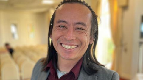 A close up of Deputy David Goy smiling. He has long dark hair and is wearing a red shirt with a grey jacket.