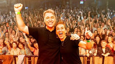 Jonny Owen and Vicky McClure with their arm around each other, the opposite arms aloft, as they stand on stage with a crowd of people cheering and waving behind them