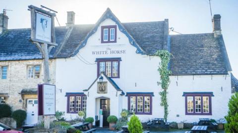 The White Horse Inn from the front. It is a white building with red window frames and picnic benches outside.