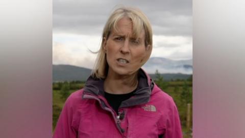 Katie Hunter stands outside wearing a pink windproof coat, gesturing as she speaks to camera in front of heathland and cloudy sky in near Aviemore, Scotland on Saturday.