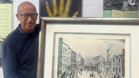 Auctioneer David Fergus, who is wearing glasses and a navy blue jumper, is stood next to a large, framed LS Lowry print. There are other paintings in the background.