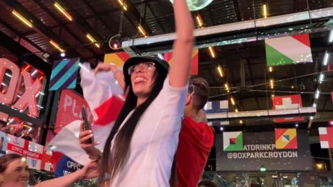 A woman wearing an England shirt and bucket hat waving her arm in the air and sticking out her tongue in celebration