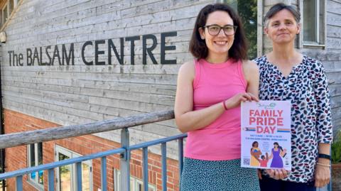 Izzie Koch and Emma Brown. Izzie has short dark hair, glasses, a pink t-shirt and a spotty skirt. She is holding a poster advertising Family Pride Day. She is smiling at the camera. Emma Brown is stood next to her. She has short grey hair and is wearing a white shirt with a blue and red pattern. She is also smiling at the camera