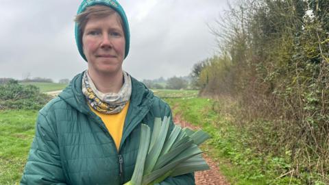 Ella Gent stand on a rural path in a field. She person is wearing a quilted green jacket, a yellow shirt underneath, a patterned neck scarf and a teal knitted hat. She is holding a bundle of long green leaves that appear to be freshly harvested leeks.
