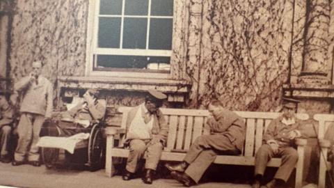 Five World War One soldiers, dressed in hospital uniforms, relax on a bench in front of a mansion. One man has an arm in a sling. Another man is in a wheelchair.