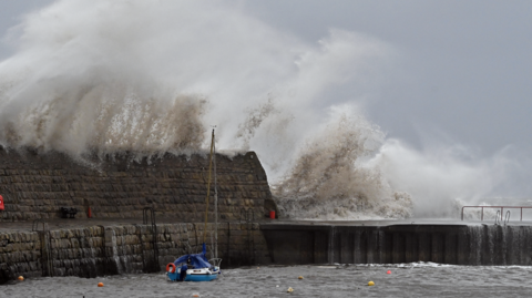 Large waves crashing over a sea wall, dwarfing a small fishing in a harbour