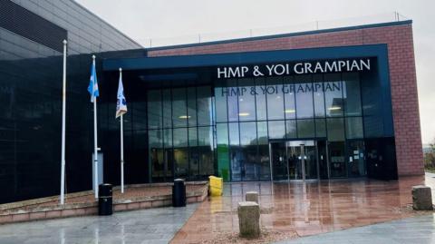 Brick and glass building with sign wording saying 'HMP & YOI Grampian', and flagpoles at the side.