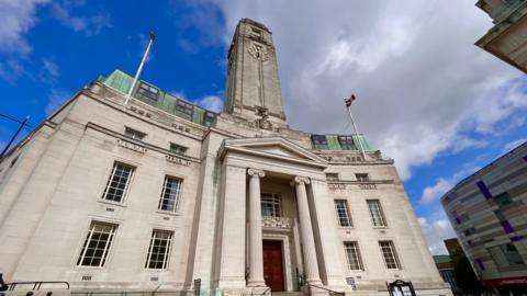 A picture of Luton Council Headquarters in Luton. It's a grey-ish white building. It has a neoclassical style with art deco detailing and the sky is blue behind it aside from one greyish cloud.