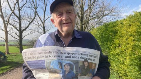 Alan Lawty pictured standing next to a hedge holding up a newspaper. He has grey hair and is wearing a blue jumper and cap.