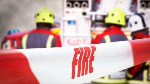 Scene of incident is cordoned off with red and white tape, reading ‘fire’. Out of focus in the background are three firefighters, two fire engine appliances and smoke billowing through the air. (stock image)