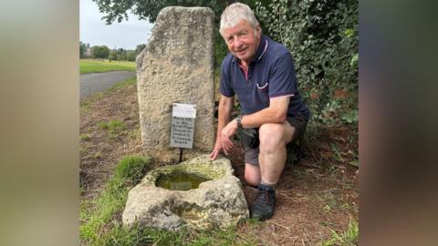 Ian Tomlinson kneels down in the grass beside two large stones; one is positioned upright and the other is basin-shaped, and filled with greenish coloured water. Ian is an older gentleman with short white hair, with a navy blue polo, brown shorts and black walking boots.