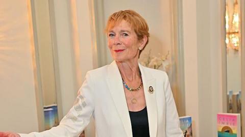 Actress Celia Imrie stood in front of a pile of her books. She is wearing a white jacket and black shirt. Her first arm is slightly raised and she is pointing her index finger.