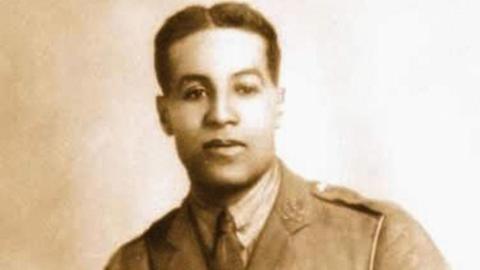 A sepia photograph of Walter Tull wearing a military jacket, tie and white shirt. His hair is combed with a parting in the middle. He is looking directly at the camera.