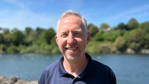 Mark Bowden smiling at the camera. He is stood in front of a reservoir with a bank with trees and grass behind it.