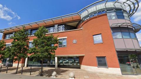 The exterior of Worcester Magistrates' Court set against blue skies on a sunny day. It's a modern building made of red brick, with curved glass at one end. Leafy trees stand on the street in front of the building.