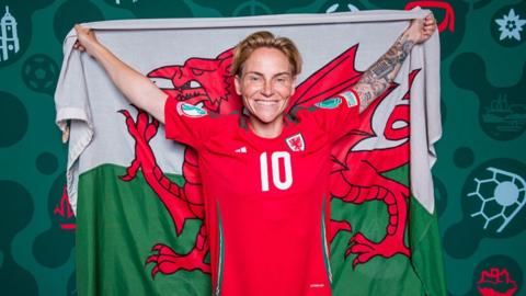 The image shows Jess Fishlock holding up the flag of Wales with a bold red dragon stretched across green and white stripes. She is wearing a red football jersey with the number 10 on the back and the Welsh national team crest. Her left arm is tattooed, and she is surrounded by a vivid green backdrop decorated with football-related symbols: a football, a lighthouse, and other stylized graphics.