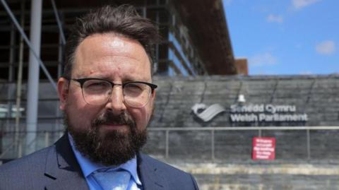 A picture of Hefin David, who is wearing a suit, has black-rimmed glasses, dark hair and a dark beard, stood in front of the Senedd building.