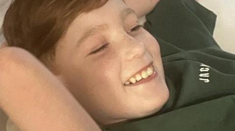 Archie is wearing a green t-shirt, has red hair and is lying in a hospital bed smiling at the camera.