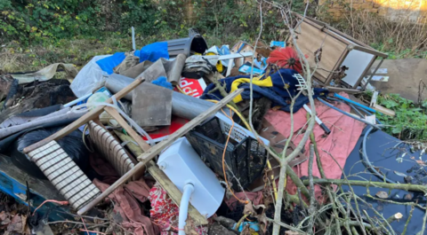 A big pile of rubbish in a wooded area including chairs and other furniture.
