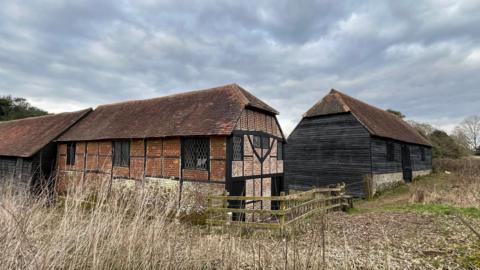 An exterior view of a group of Tudor barns, with brown brickwork, a slate roof and black oak beams.