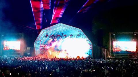 A picture of the stage at Live in Somerset.  The photo was taken in the dark and the stage is lit up in different colours.  There are two big screens either side of the stage