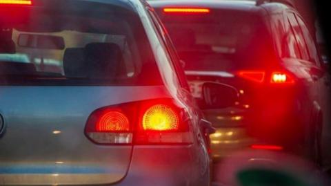 An unidentified silver car in a traffic queue with its rear brake lights illuminated red