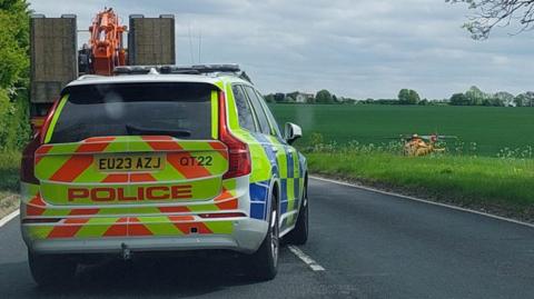 A four-by-four police car parked on the single-carriageway road. A red and yellow air ambulance has landed on a large field to the right of the image.