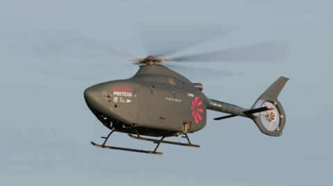 A helicopter without a window/cockpit flying in the blue sky. It is dark grey with the name Proteus on the front side in white and a pink logo of a rotor blade printed at the back 