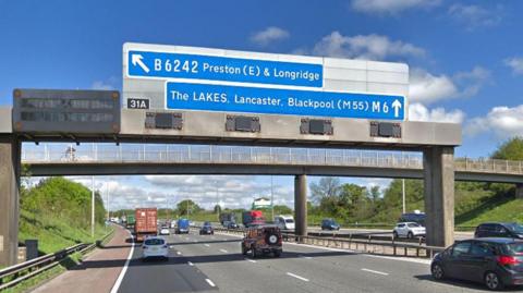 Sign over the M6 motorway near Preston pointing north to the Lakes, Lancaster and Blackpool and west towards Preston and Longridge at junction 31A. Cars are driving underneath it