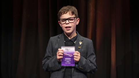 Frazer Tibbitts, who has short brown hair and black framed glasses, stands in front of a dark brown material backdrop. He is wearing a black polo neck and dark grey blazer and holding up a purple Make a Difference Award with both hands