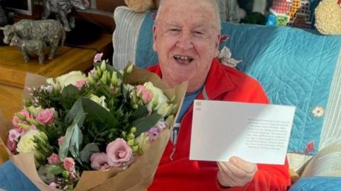 A man wearing a red top and shorts, sits on a chair while holding a bouquet of flowers in one hand and a note in the other.