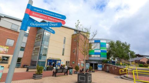 The outside of the University of North Durham Hospital. It a brown and beige-coloured brick building. The entrance has green, white and blue rectangle panelling. There is a sign pointing patients to various departments.  A small tree is in a plant pot in a seating area.