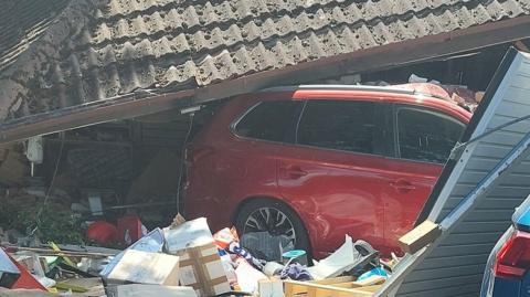Part of a red car can be seen crashed through a garage. The tiled roof of the building is on top of the car and a battered steel garage door can be seen off its hinges. There are boxes and other items that were inside the garage around the wheels of the car. 