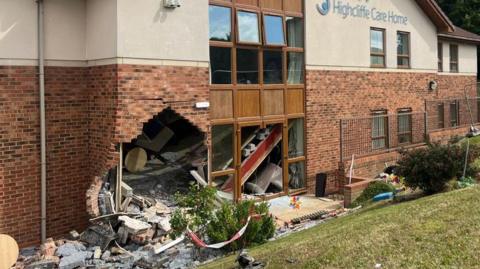 A large hole at the bottom left of a brick and white stone building next to a window. Bricks and debris from the wall are scattered all around and in the bushes and grass in front of the building.