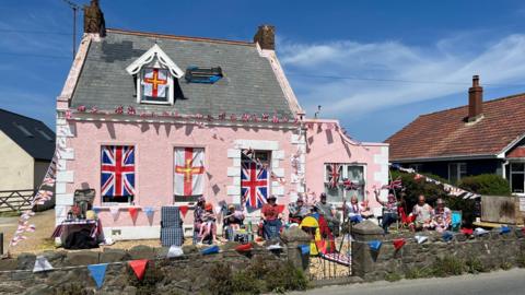 A pink cottage covered in Guernsey and Union flags in the sunshine, with people sitting outside.