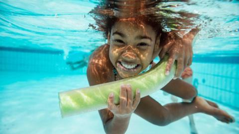 A child is smiling whilst holding a pool buoyancy aid underwater in a swimming pool.