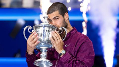 Carlos Alcaraz, wearing a purple Nike jacket, kissing the US Open trophy.