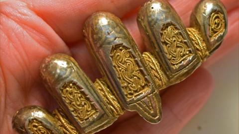 A close-up of a Viking-era silver and gold sword pommel resting on a hand. It is made up of five lobes, which decrease in width from the tallest on in the middle. Each of the lobes has an inset panel of with an interlocking pattern of gold wire on a gold panel. More gold wire is woven between the lobes.