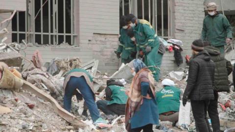 About six people in green jackets look through the rubble outside a building. The remains of two windows with vertical white bars across the holes can be seen. Another three men are looking on and a woman walks past. 